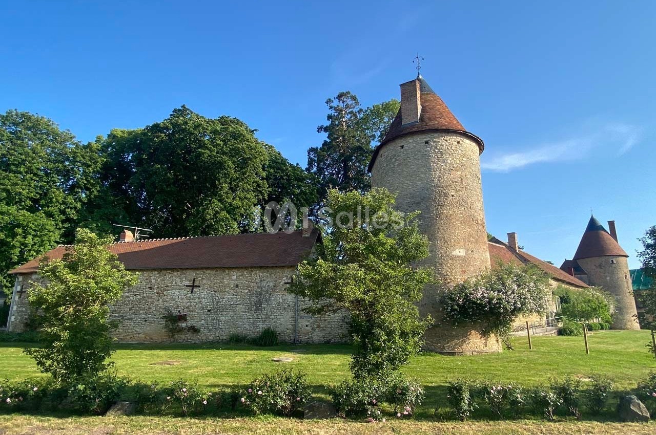 Bâtiment en pierre avec tours rondes et toits en tuiles, entouré d'arbres et d'une pelouse verdoyante.