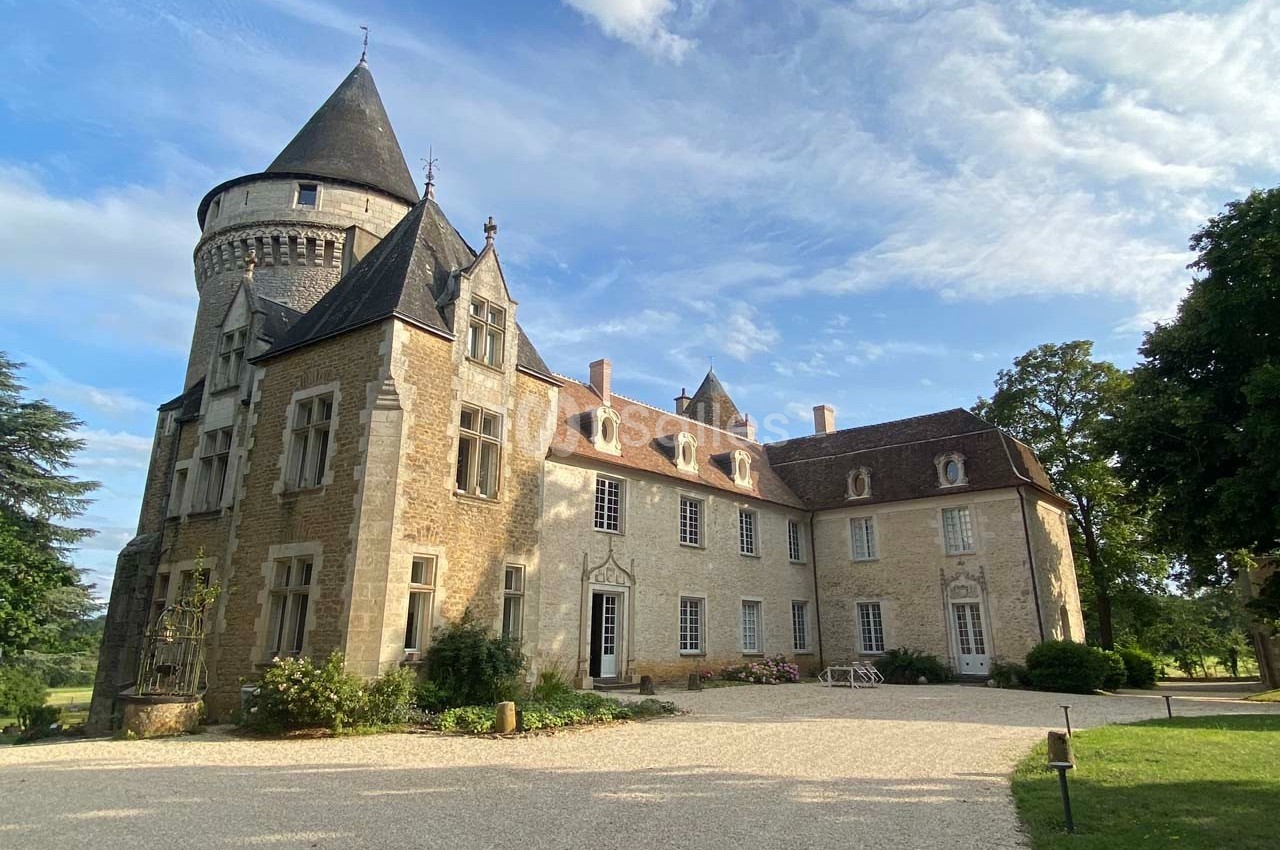 Château en pierre avec tour ronde, entouré d'arbres, sous un ciel partiellement nuageux.