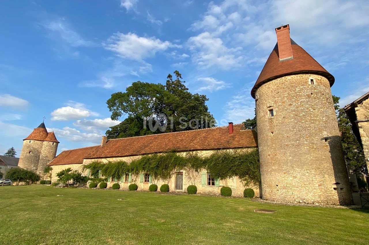 Bâtiment en pierre avec tours rondes et toit en tuiles rouges, entouré d'une pelouse et d'arbres sous un ciel bleu.