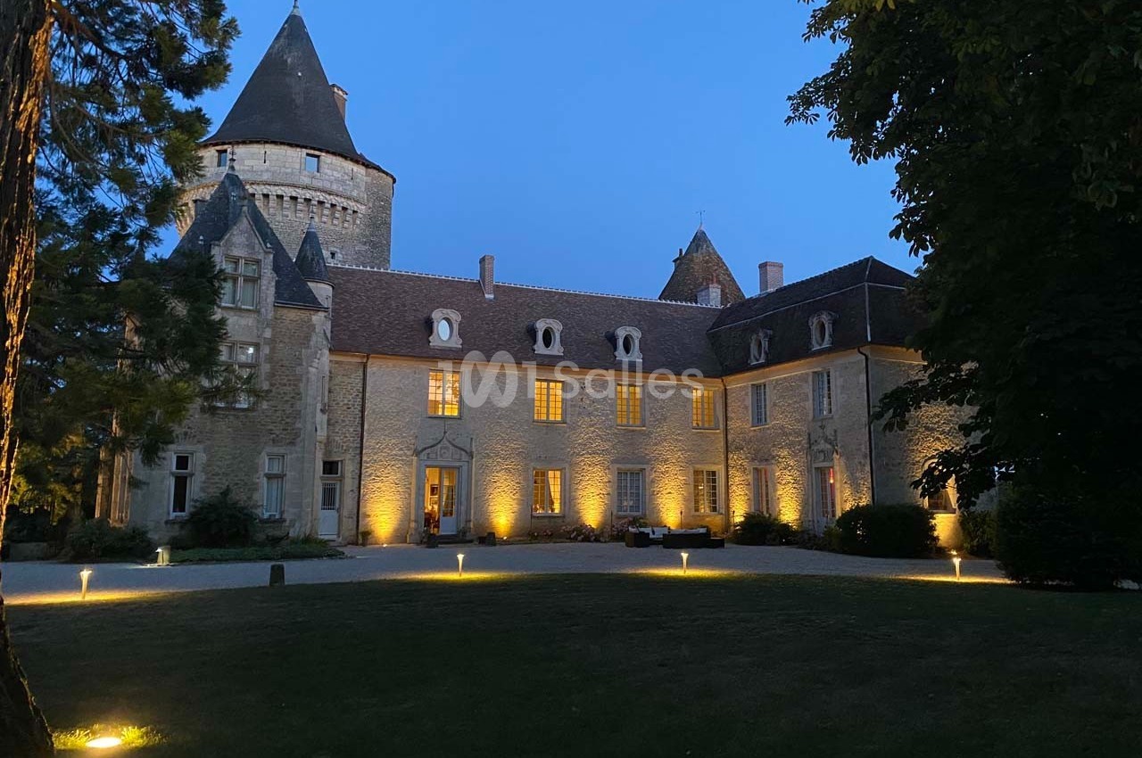Façade d'un château en pierre éclairé par des lumières au crépuscule, entouré d'arbres et d'une pelouse.