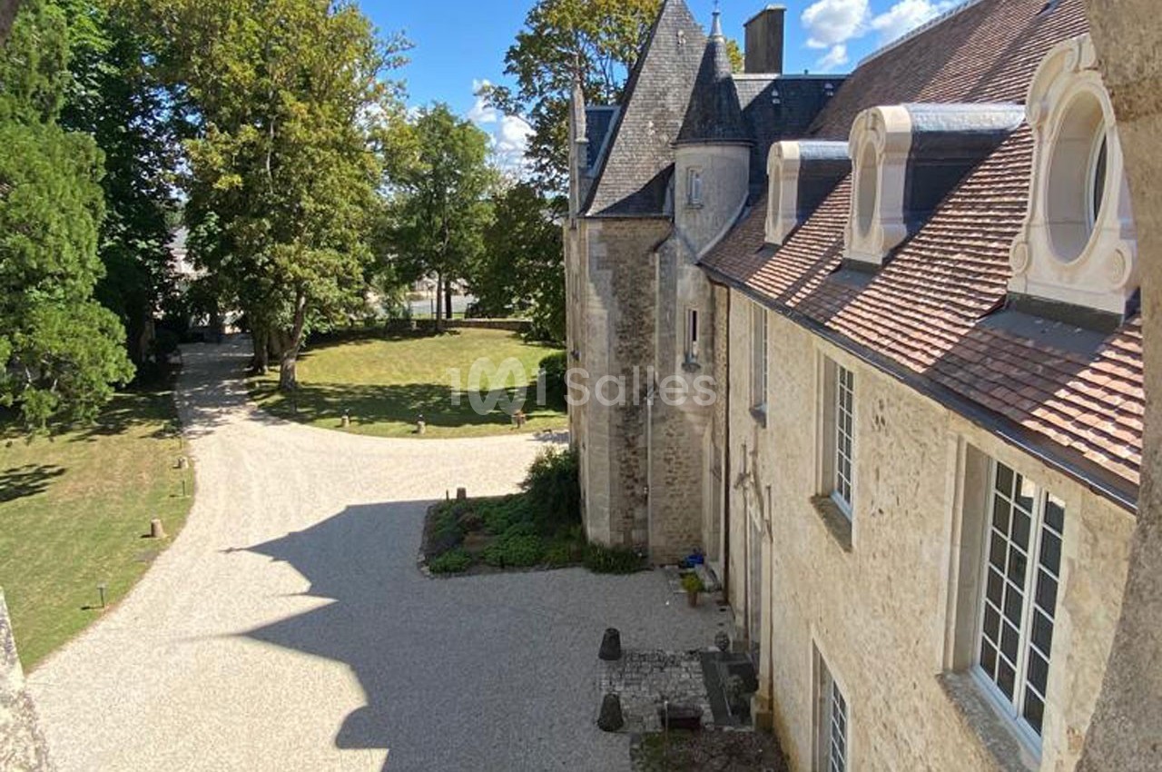 Vue d'un bâtiment historique en pierre avec des toits en ardoise, entouré d'arbres et d'une allée gravillonnée.