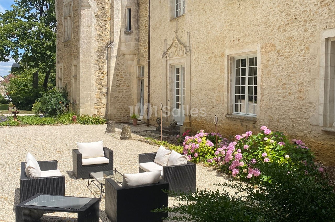 Mobilier de jardin en rotin noir avec coussins beiges, disposé sur une cour gravillonnée près d'une bâtisse en pierre.