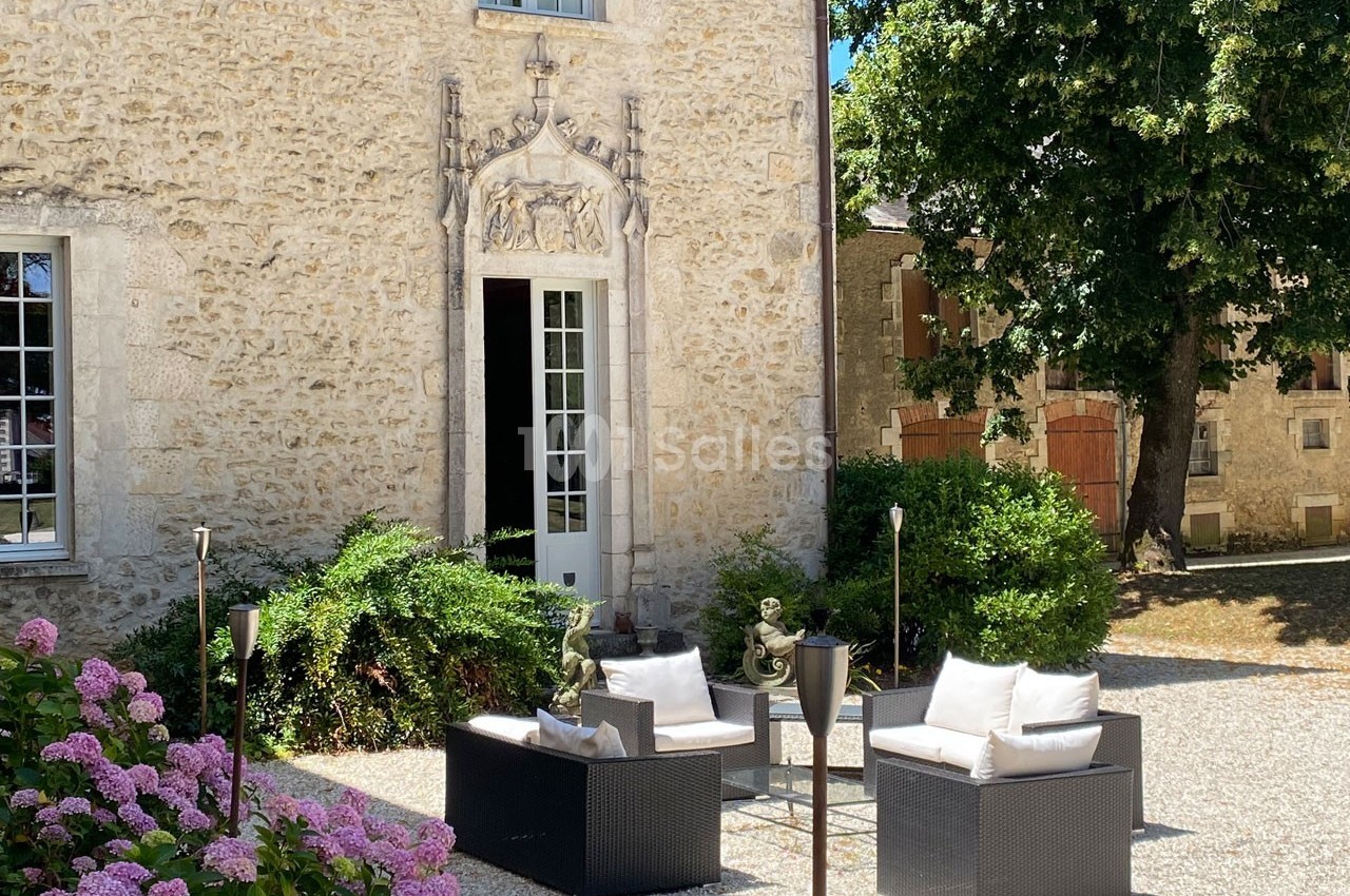 Mobilier de jardin en rotin avec coussins blancs devant une façade en pierre ornée, entourée de verdure et de fleurs.