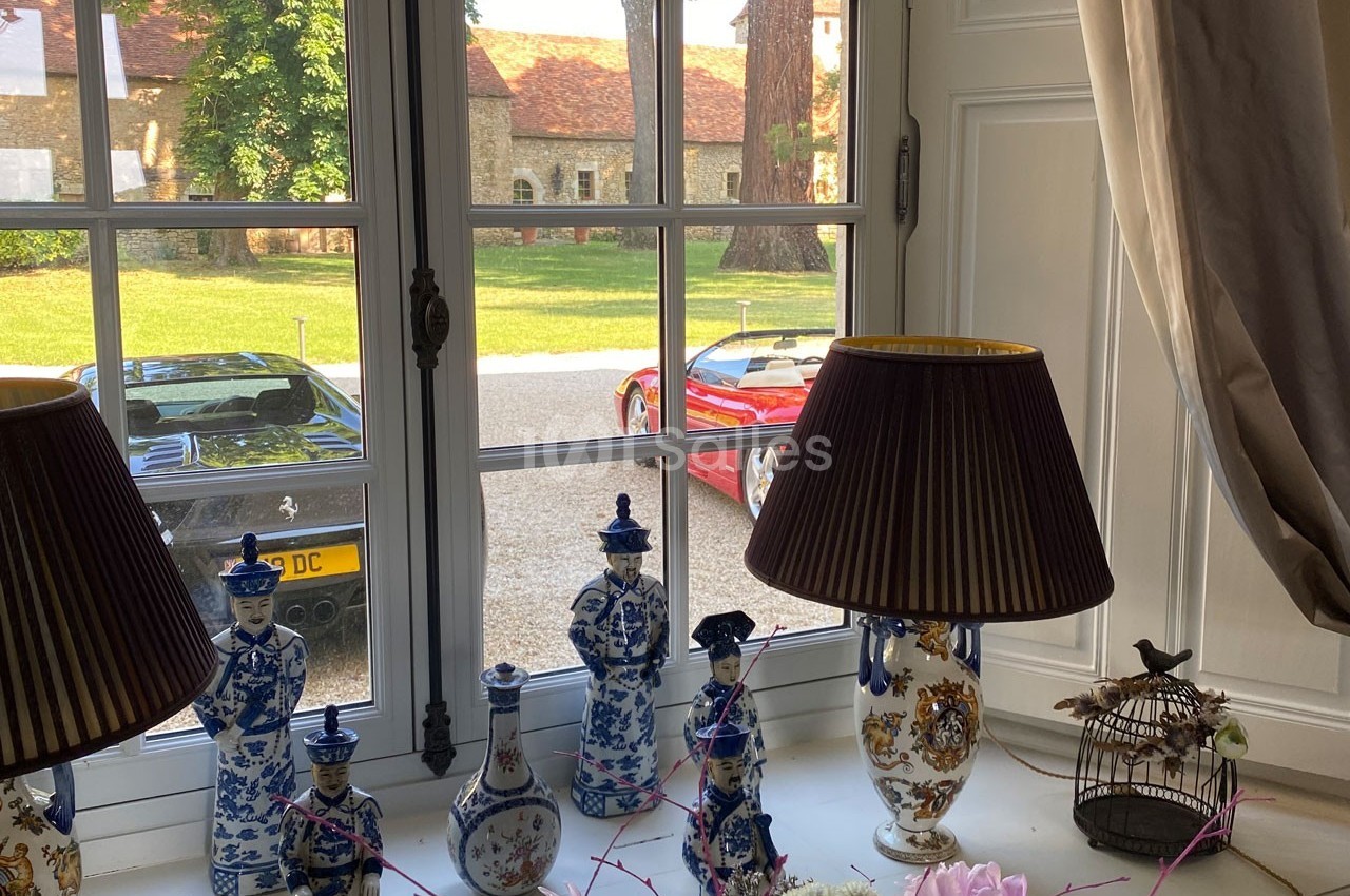 Vue depuis une fenêtre sur une cour avec deux voitures de sport, des lampes et des porcelaines décoratives au premier plan.