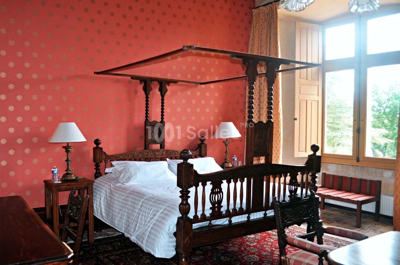Chambre avec lit à baldaquin en bois sculpté, murs rouges à motifs, grandes fenêtres et mobilier classique.