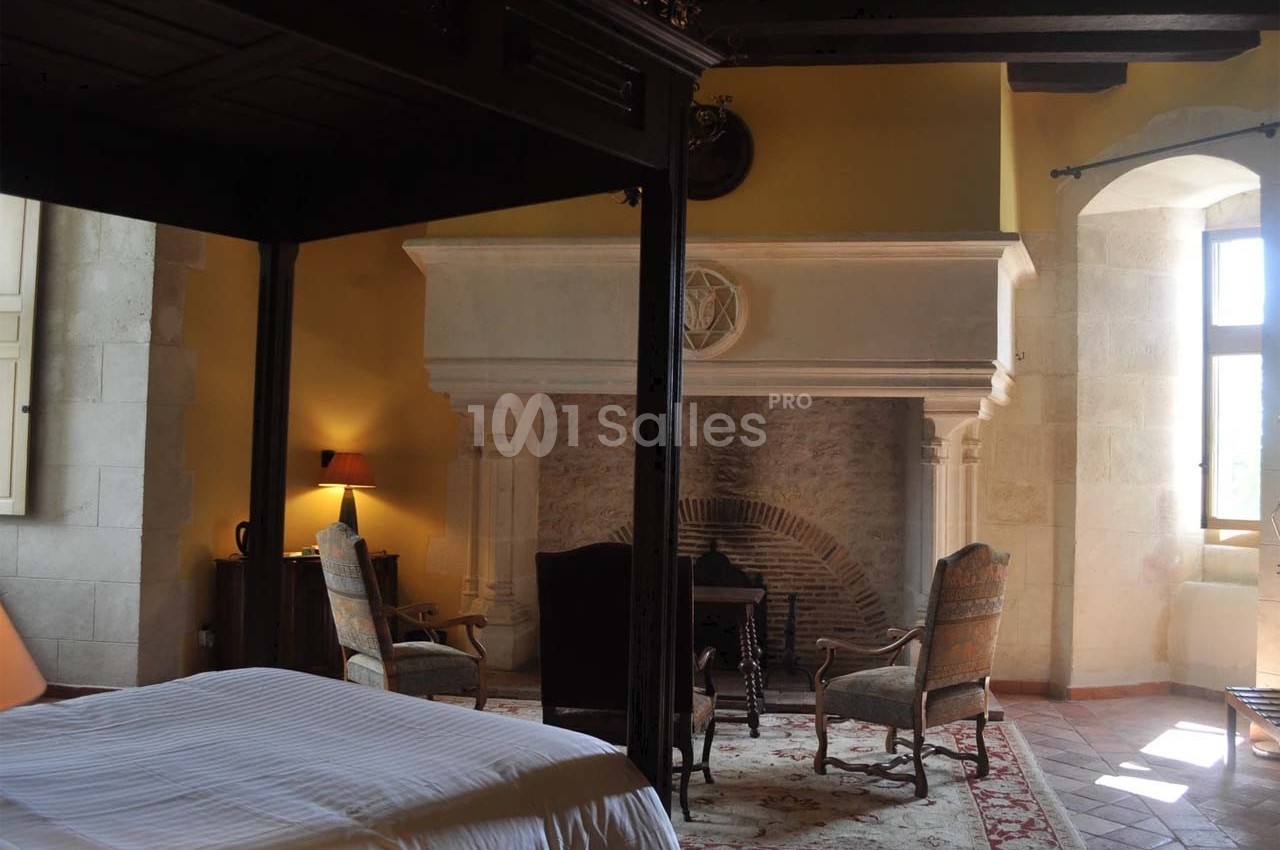 Chambre avec lit à baldaquin, cheminée en pierre, fauteuils et lumière naturelle entrant par une fenêtre.