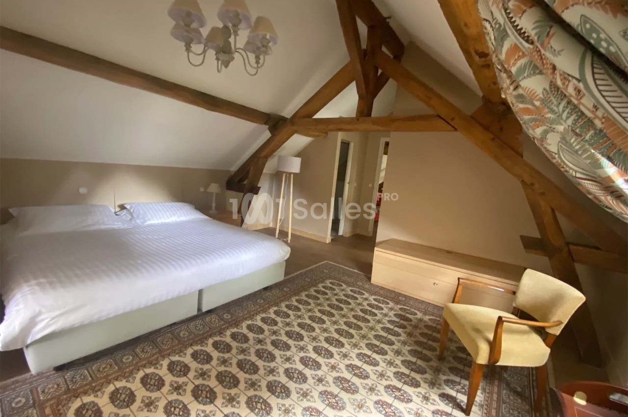 Chambre mansardée avec poutres apparentes, lit double, tapis décoratif, fauteuil et éclairage naturel.
