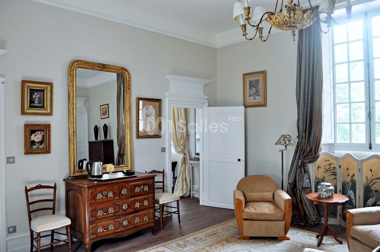 Chambre lumineuse avec mobilier classique, miroir doré, fauteuils, commode en bois et grande fenêtre à rideaux.