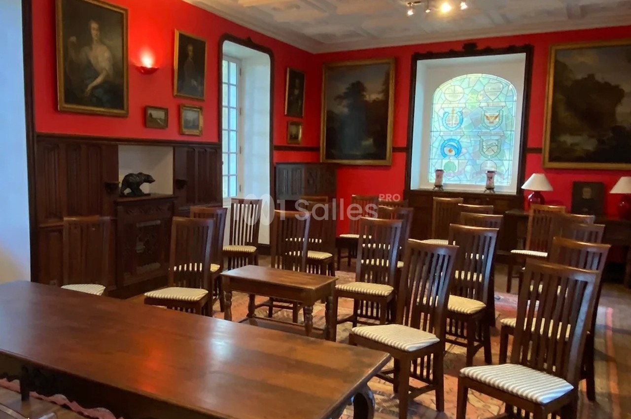 Salle avec murs rouges, chaises en bois alignées, table en premier plan, tableaux et vitraux décorant l’espace.