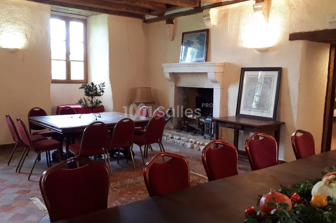 Salle avec des murs clairs, cheminée en pierre, tables en bois, chaises rouges et lumière naturelle provenant d'une fenêtre.