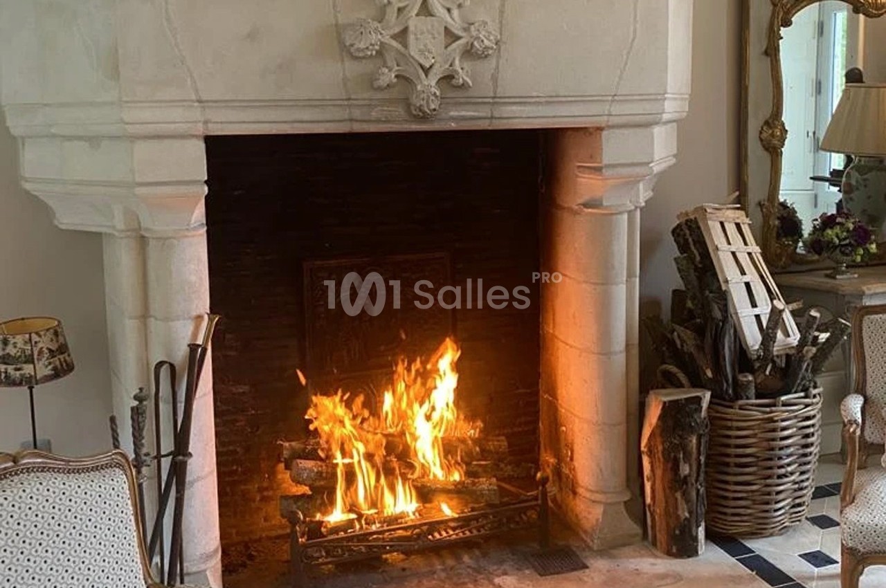 Cheminée en pierre allumée avec des bûches enflammées, entourée de fauteuils et d'un panier de bois.
