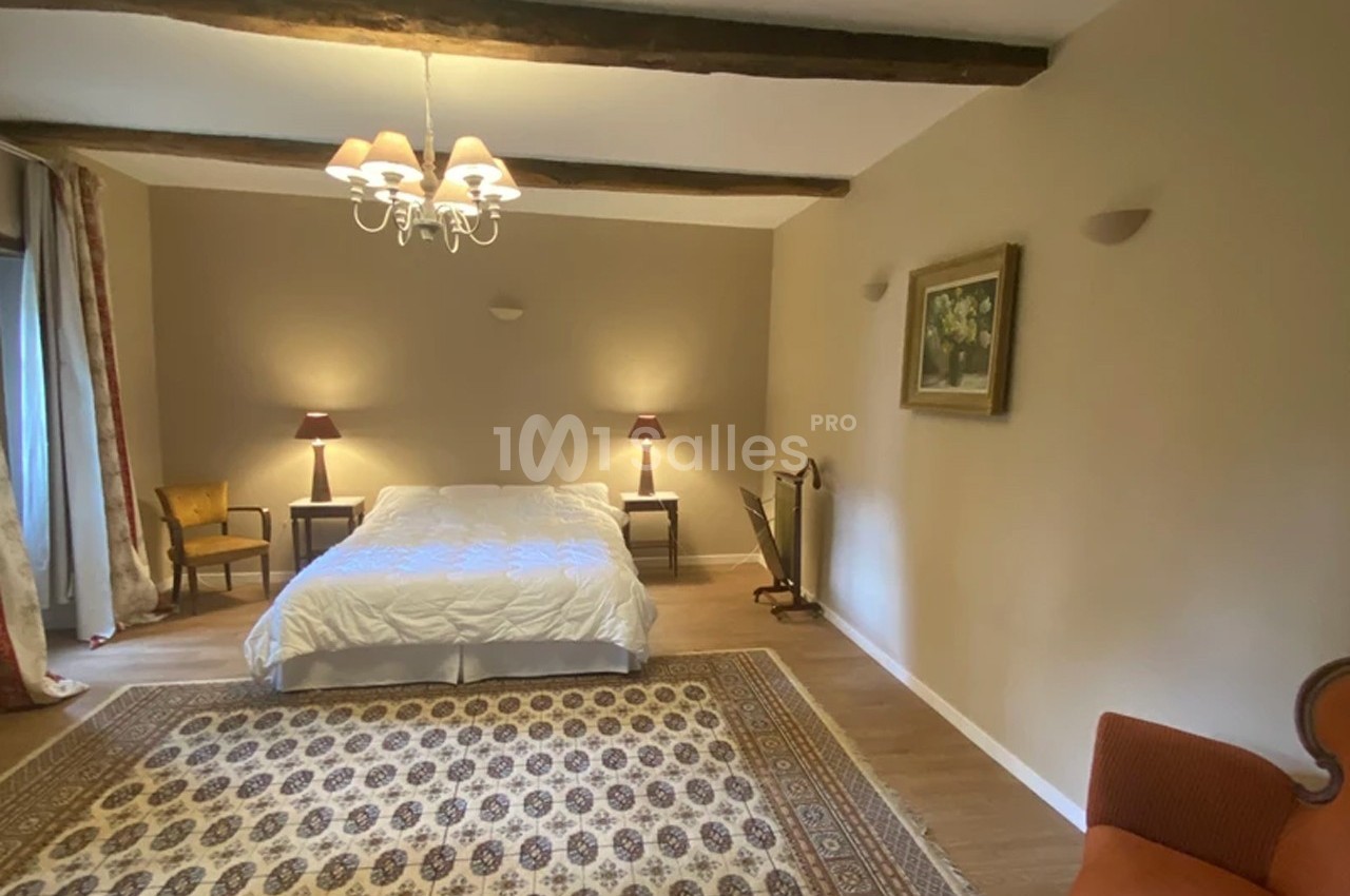 Chambre lumineuse avec lit double, tapis au sol, poutres apparentes, mobilier classique et éclairage doux.