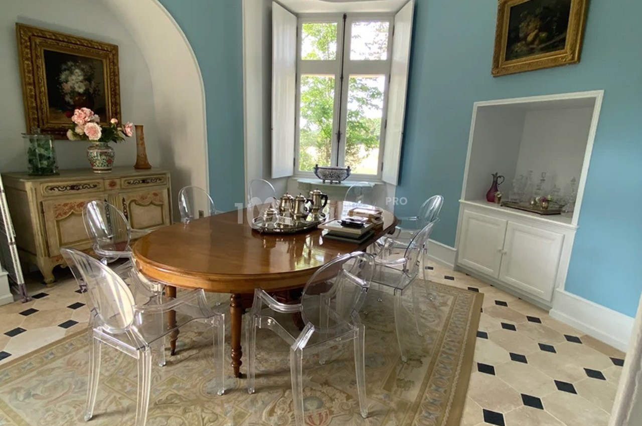 Salle à manger lumineuse avec table en bois ovale, chaises transparentes, murs bleus et vue sur un jardin par la fenêtre.