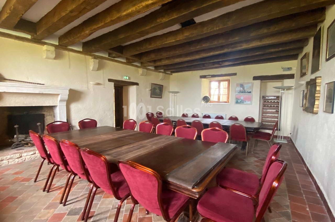 Salle avec poutres apparentes, tables en bois sombre et chaises rouges, sol en carrelage et cheminée en pierre.