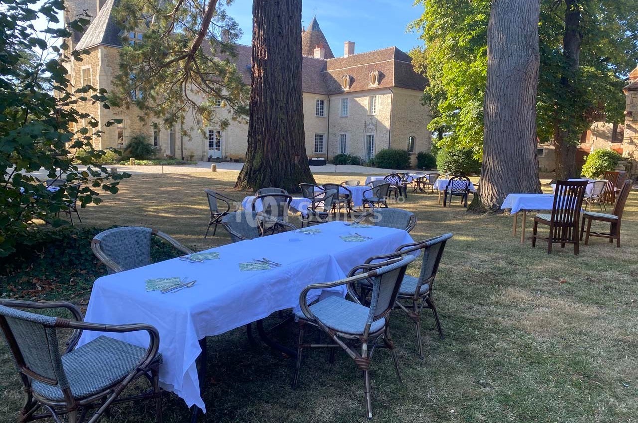 Tables et chaises en osier disposées sur une pelouse ombragée devant un bâtiment en pierre de style ancien.
