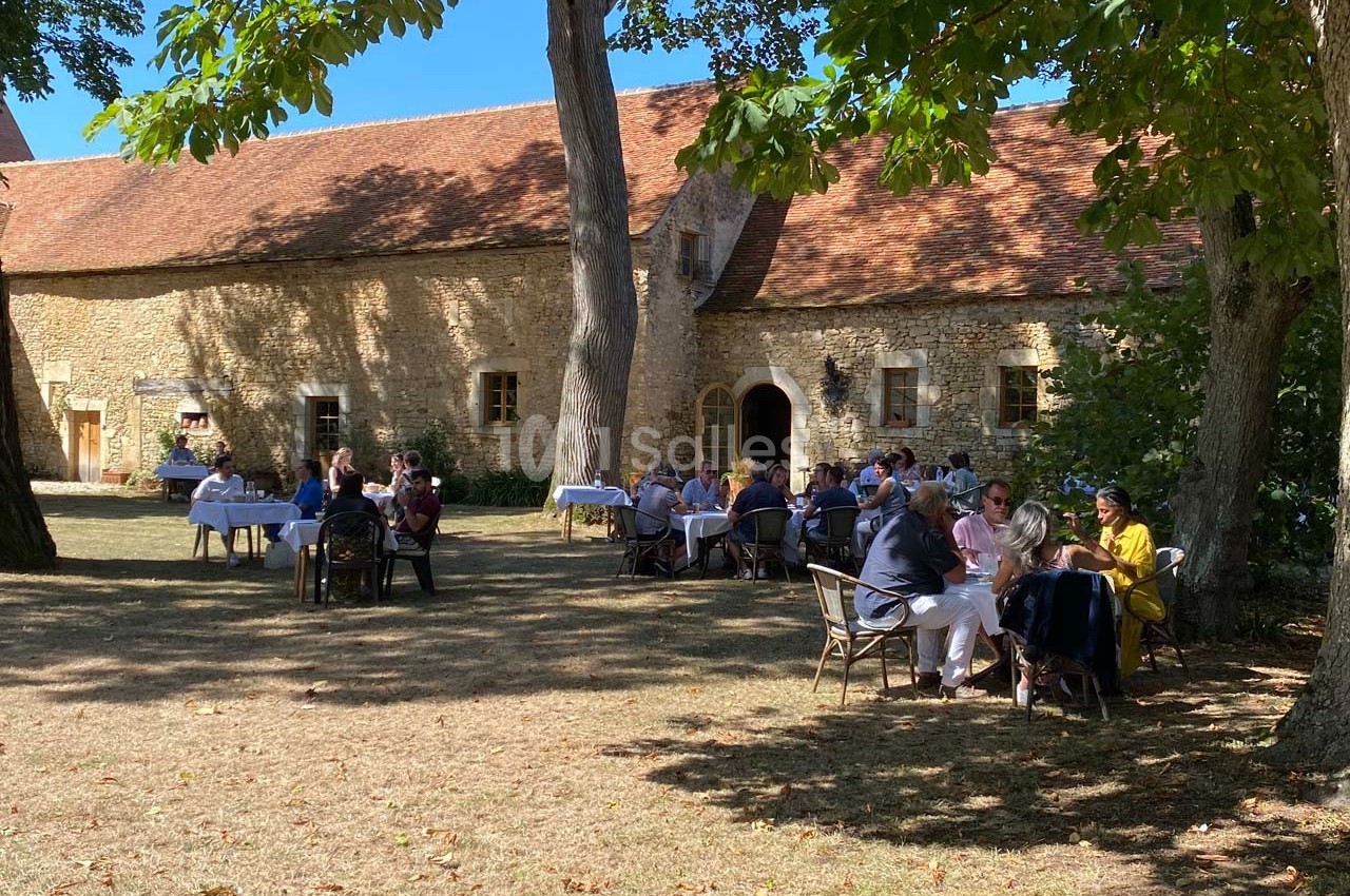 Personnes attablées dans un jardin ombragé devant un bâtiment en pierre avec toit en tuiles rouges.