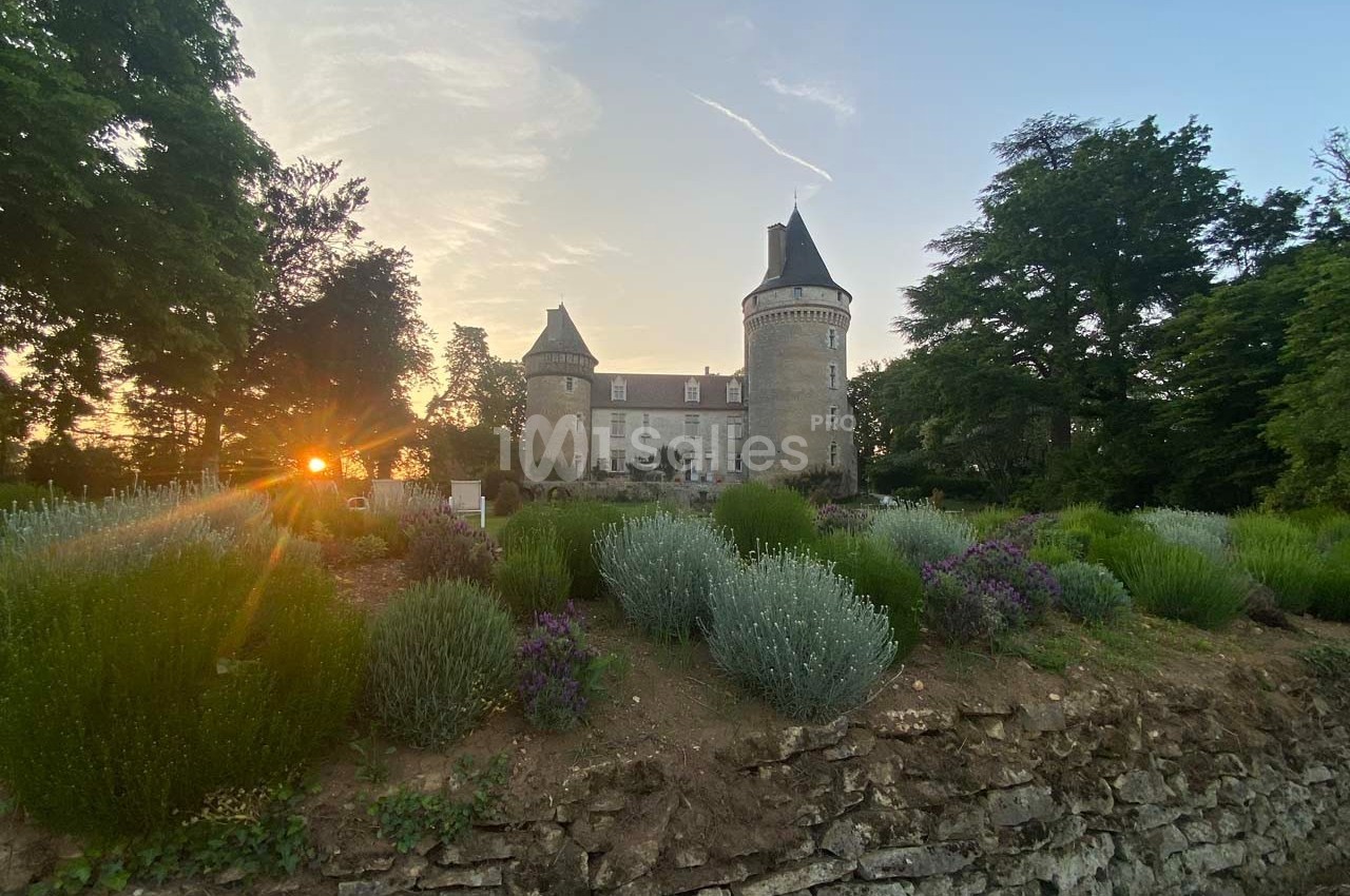 Château en pierre entouré de jardins fleuris, au coucher du soleil, avec des arbres en arrière-plan.