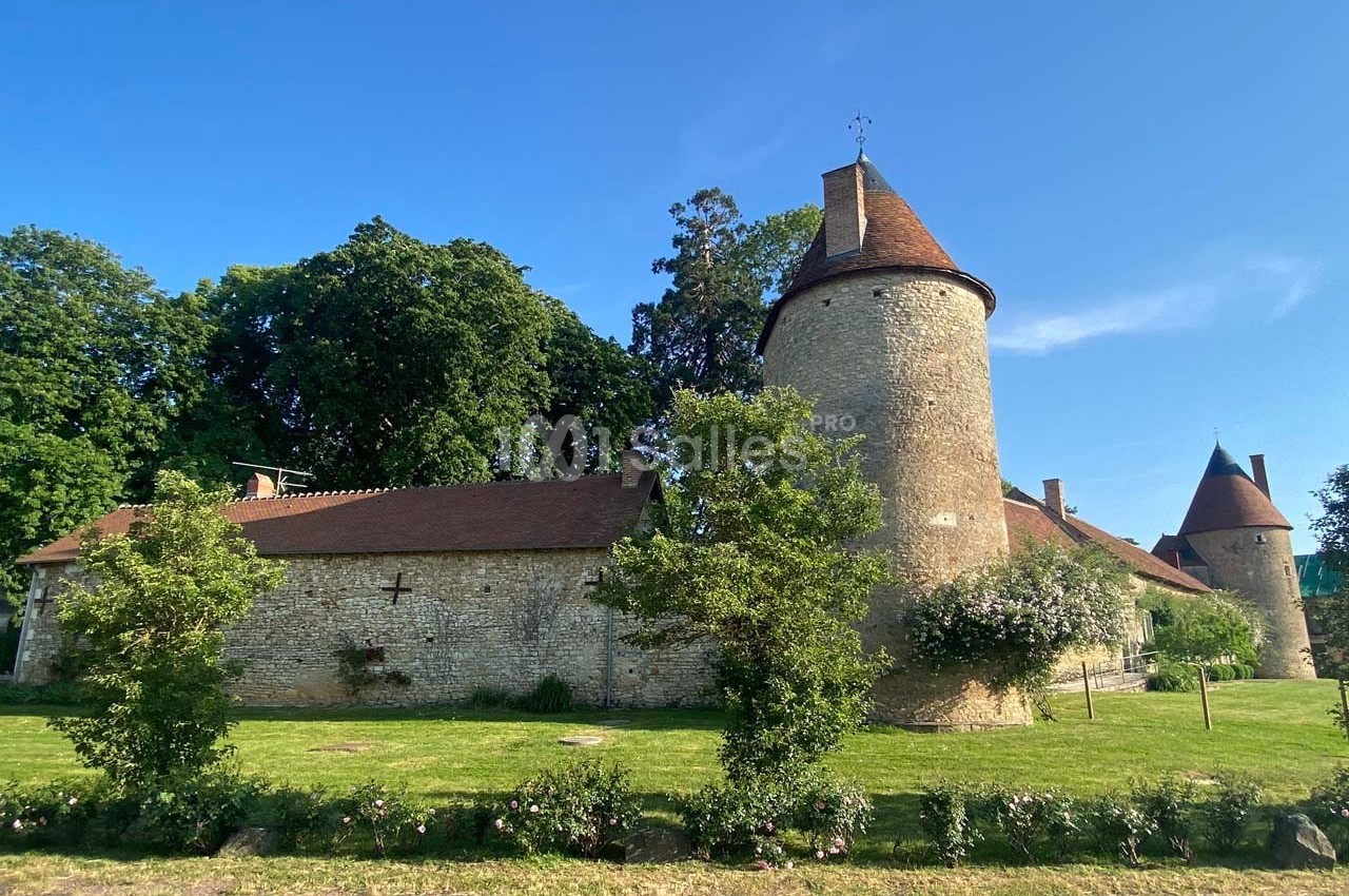 Bâtiment en pierre avec tours rondes et toits en tuiles, entouré d'arbres et d'une pelouse verdoyante.