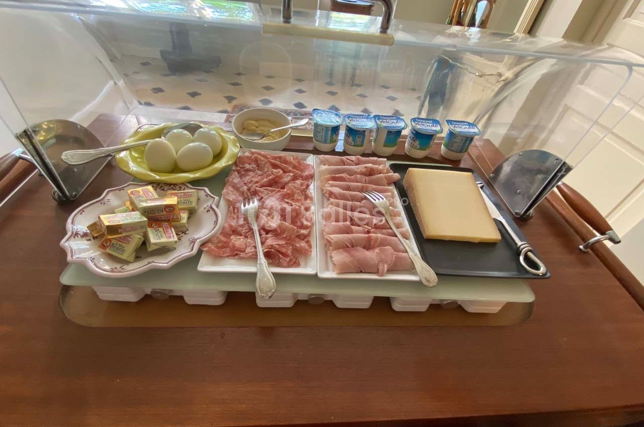 Plateau de petit-déjeuner avec charcuterie, fromage, œufs durs, beurre, yaourts et moutarde, sous une cloche transparente.