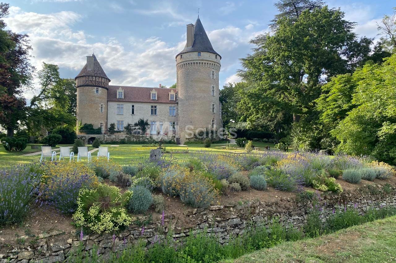 Château en pierre entouré de jardins fleuris, avec des arbres et un ciel partiellement nuageux en arrière-plan.