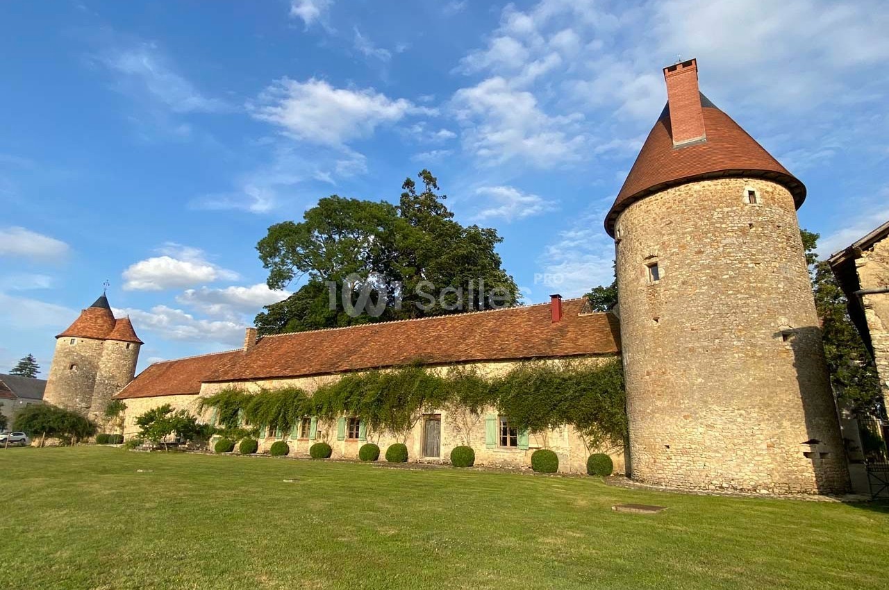Bâtiment en pierre avec tours rondes et toit en tuiles rouges, entouré d'une pelouse et d'arbres sous un ciel bleu.