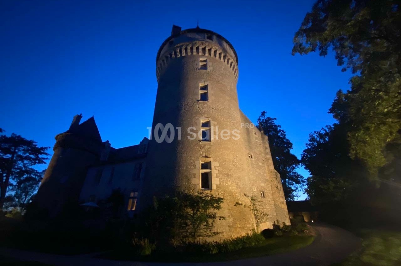 Château en pierre éclairé à la tombée de la nuit, entouré d'arbres et d'un chemin sinueux.