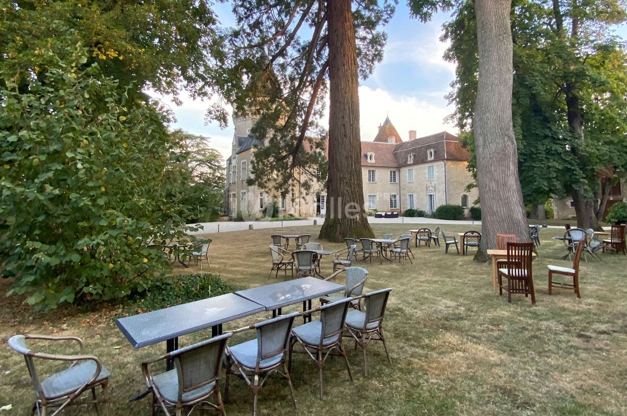 Tables et chaises disposées sur une pelouse ombragée devant un bâtiment historique entouré d'arbres.