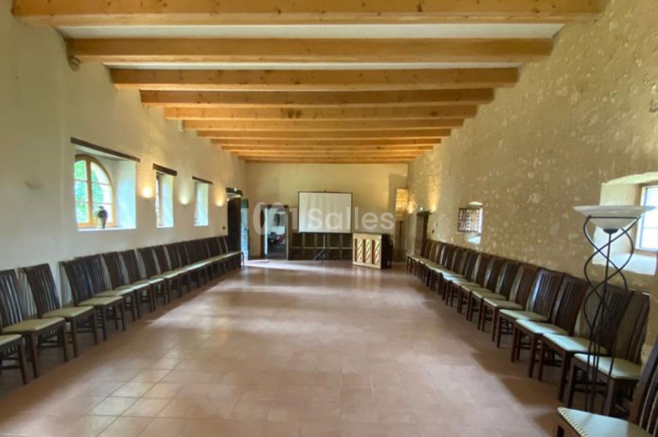 Salle lumineuse avec murs en pierre, poutres apparentes, chaises alignées le long des murs et écran de projection au fond.