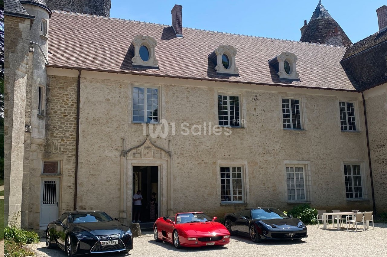 Façade d'un bâtiment ancien en pierre avec trois voitures de sport garées devant sur une cour pavée.