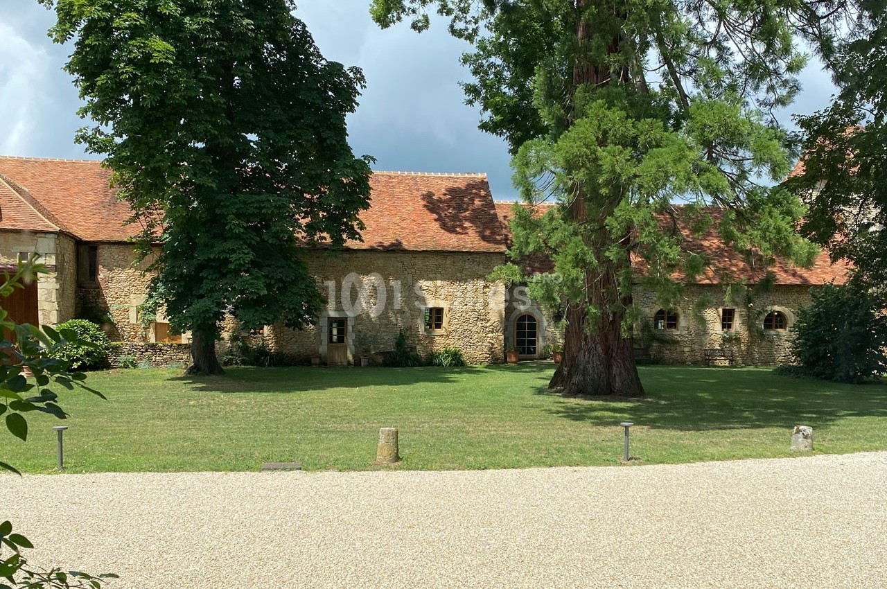 Bâtiment en pierre avec toit en tuiles rouges, entouré d'arbres et d'une pelouse, sous un ciel partiellement nuageux.