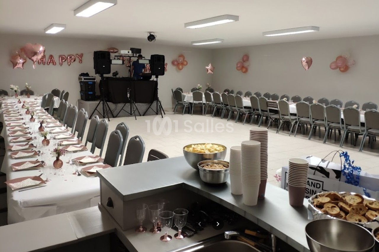 Salle décorée pour une fête avec tables dressées, chaises alignées, ballons roses et espace pour un DJ.