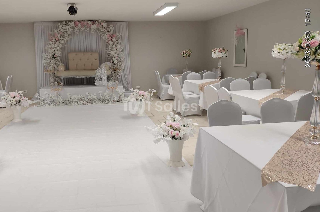 Salle décorée pour un événement avec arche florale, chaises blanches, nappes et arrangements floraux élégants.