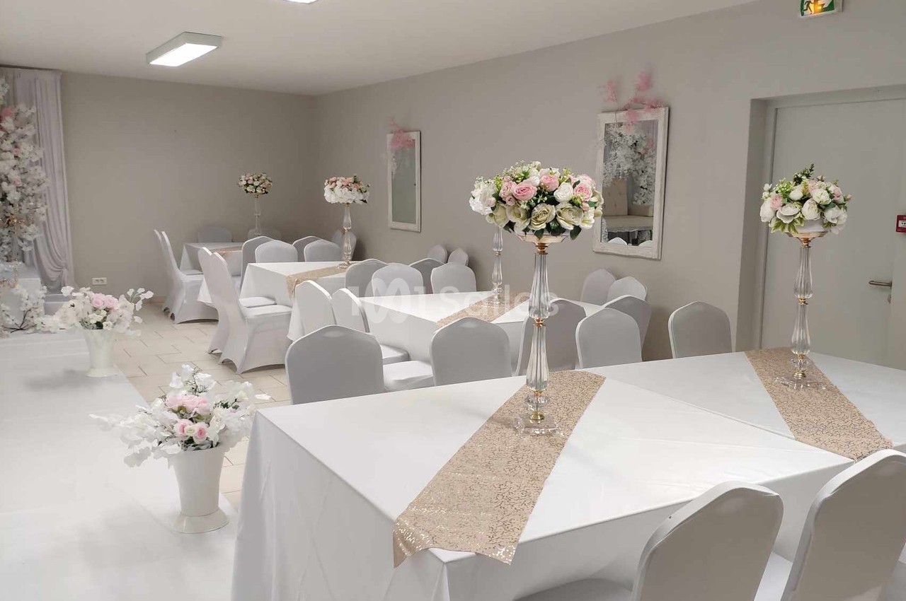 Salle de réception décorée avec des tables blanches, chemins de table beiges et centres de table fleuris.