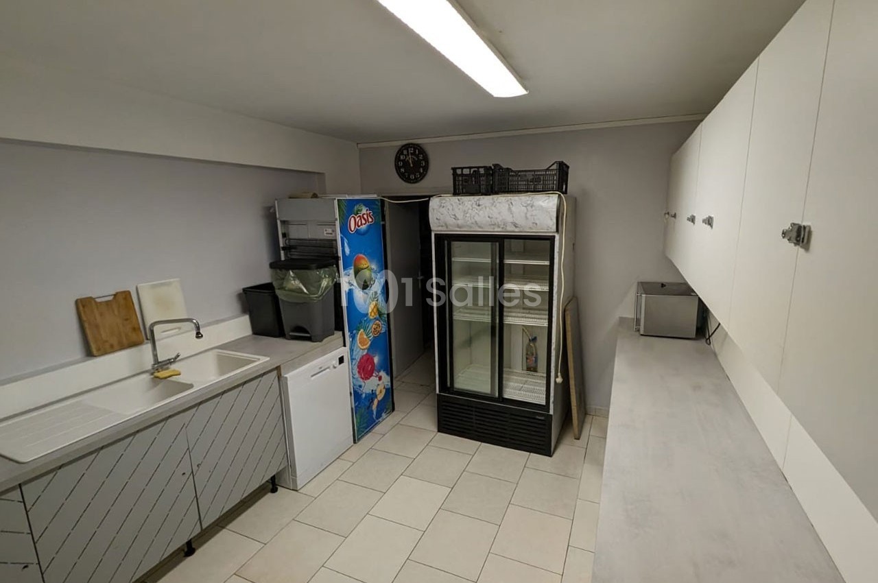 Cuisine fonctionnelle avec évier, réfrigérateurs, rangements muraux et sol carrelé dans un espace sobre et éclairé.