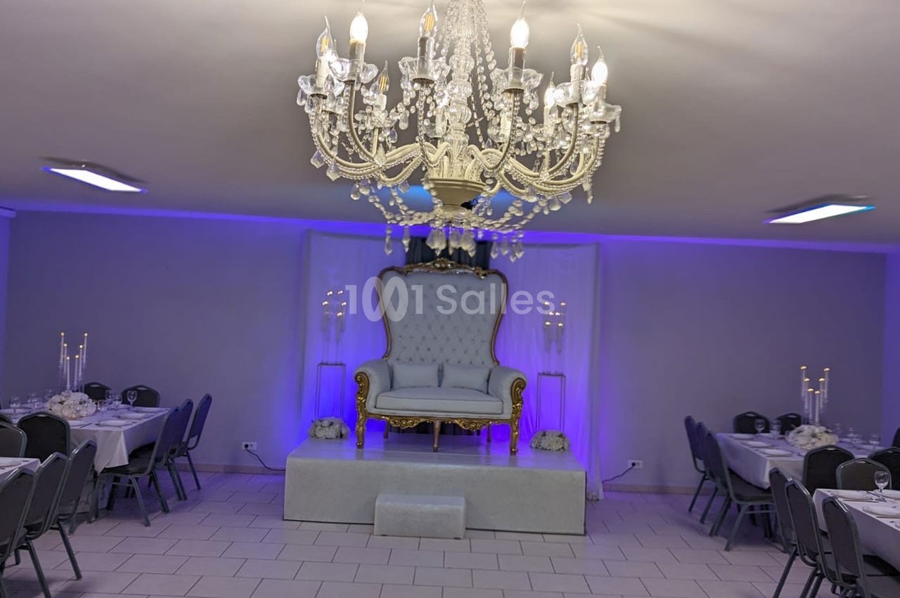 Salle de réception décorée avec un grand lustre, un fauteuil élégant sur une estrade et des tables dressées.