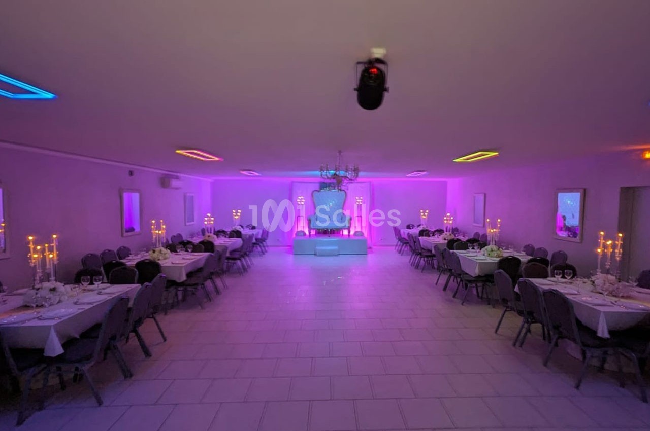 Salle de réception décorée avec des tables dressées, éclairage coloré et chandeliers allumés.