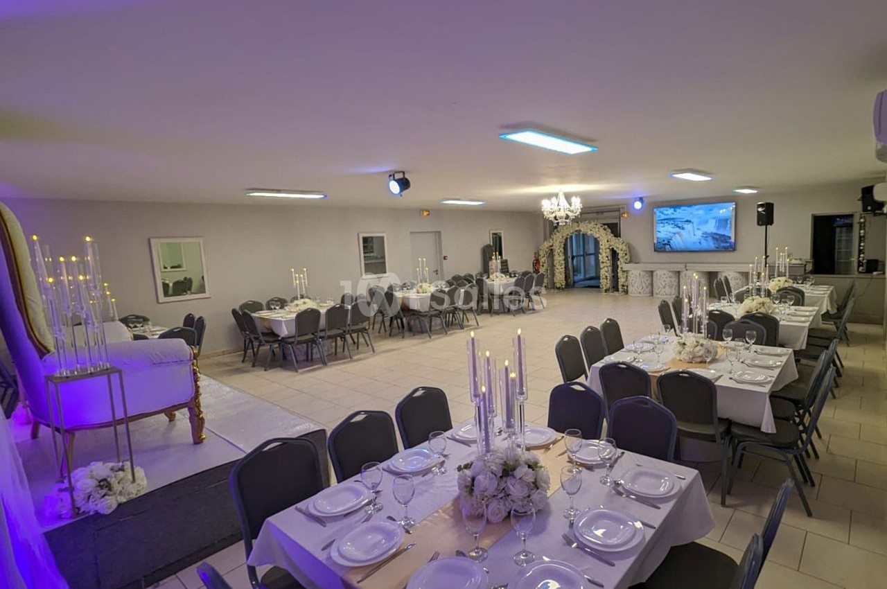 Salle de réception décorée avec des tables dressées, des chandeliers et une estrade avec fauteuil, éclairage tamisé.