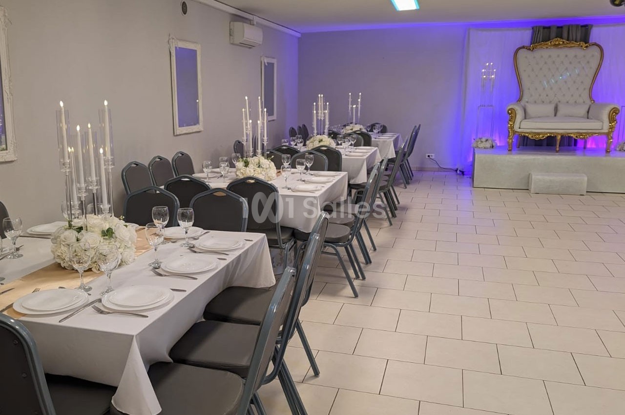 Salle de réception décorée avec des tables dressées, chandeliers et un fauteuil élégant sur une estrade.