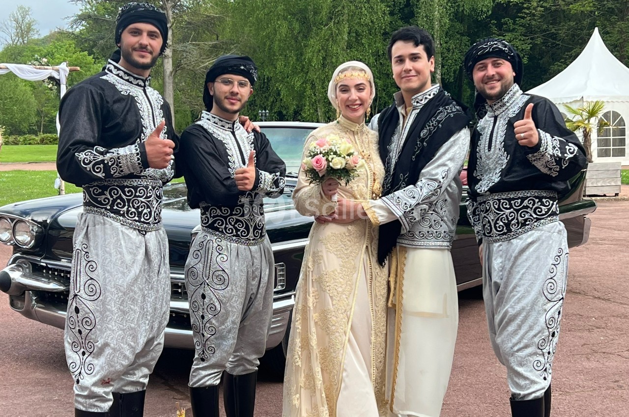 Un couple en tenue traditionnelle pose avec trois hommes vêtus de costumes similaires devant une voiture ancienne.