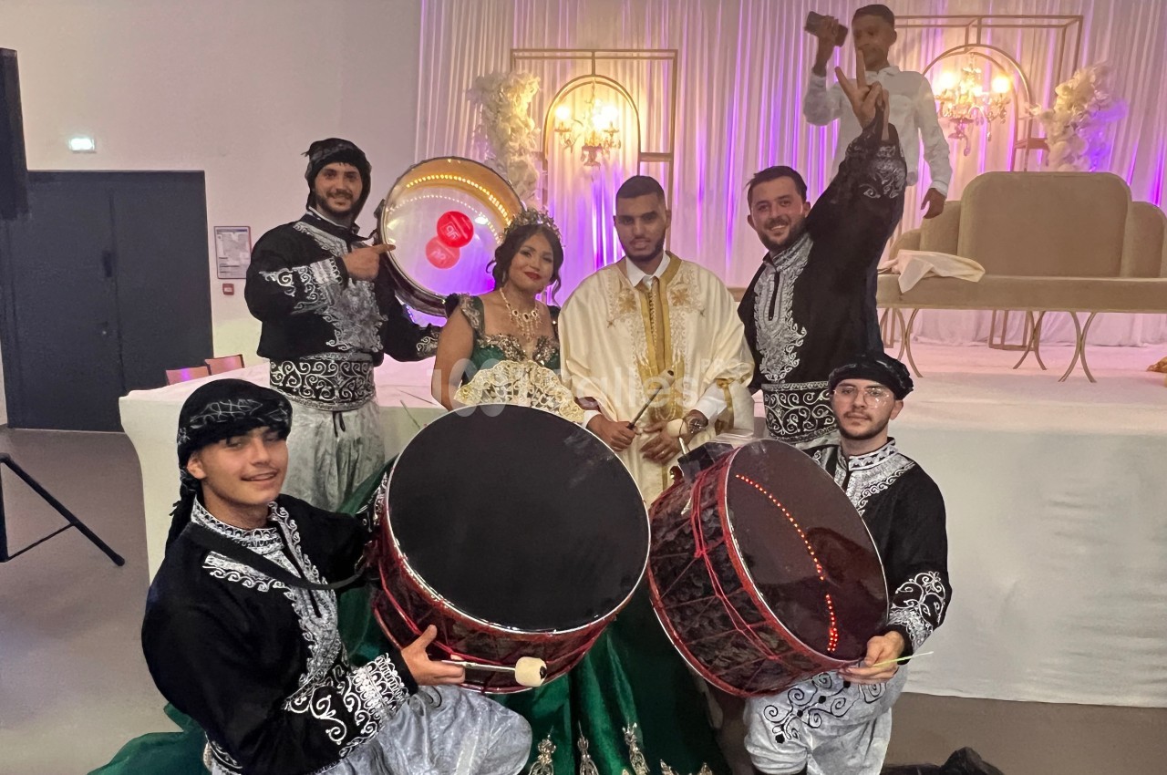 Un couple en tenue traditionnelle pose entouré de musiciens en costume jouant du tambour dans une salle décorée.