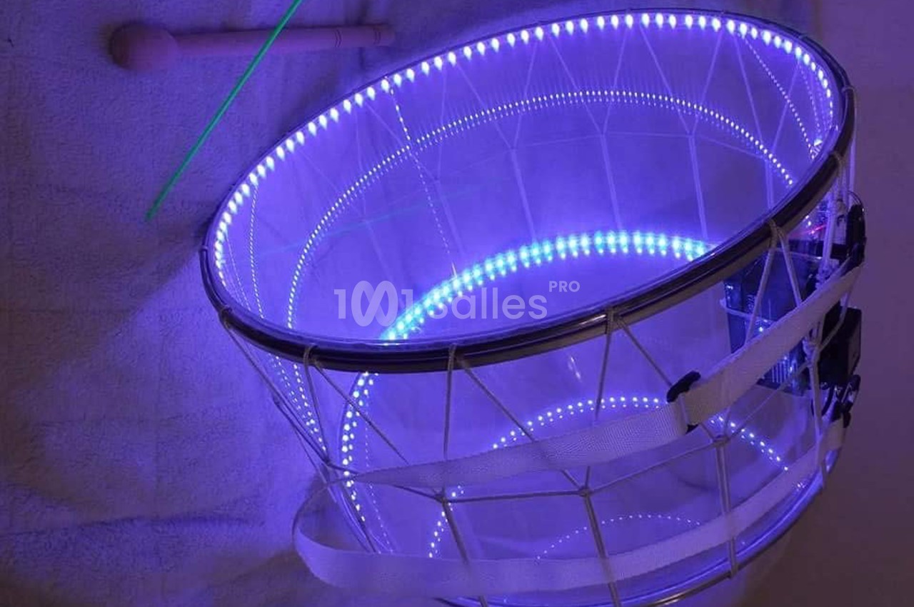 Tambour transparent éclairé par des LED violettes, posé sur une surface claire avec une baguette et une tige verte.