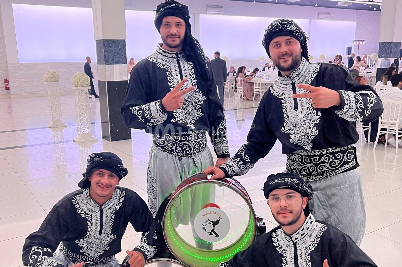 Quatre hommes en tenue traditionnelle posent autour d'un tambour décoré, dans une salle de réception lumineuse.
