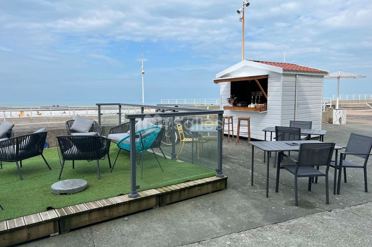 Terrasse en bord de mer avec chaises, tables, espace gazonné et une petite cabane en bois servant de kiosque.
