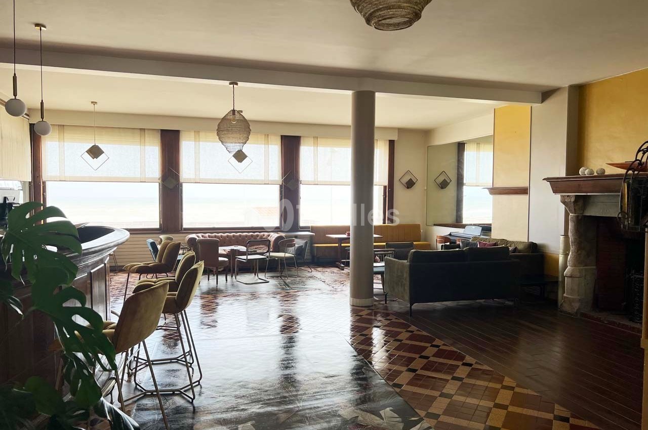Intérieur d'un salon lumineux avec des fauteuils, un bar, des fenêtres donnant sur l'extérieur et un sol en mosaïque.