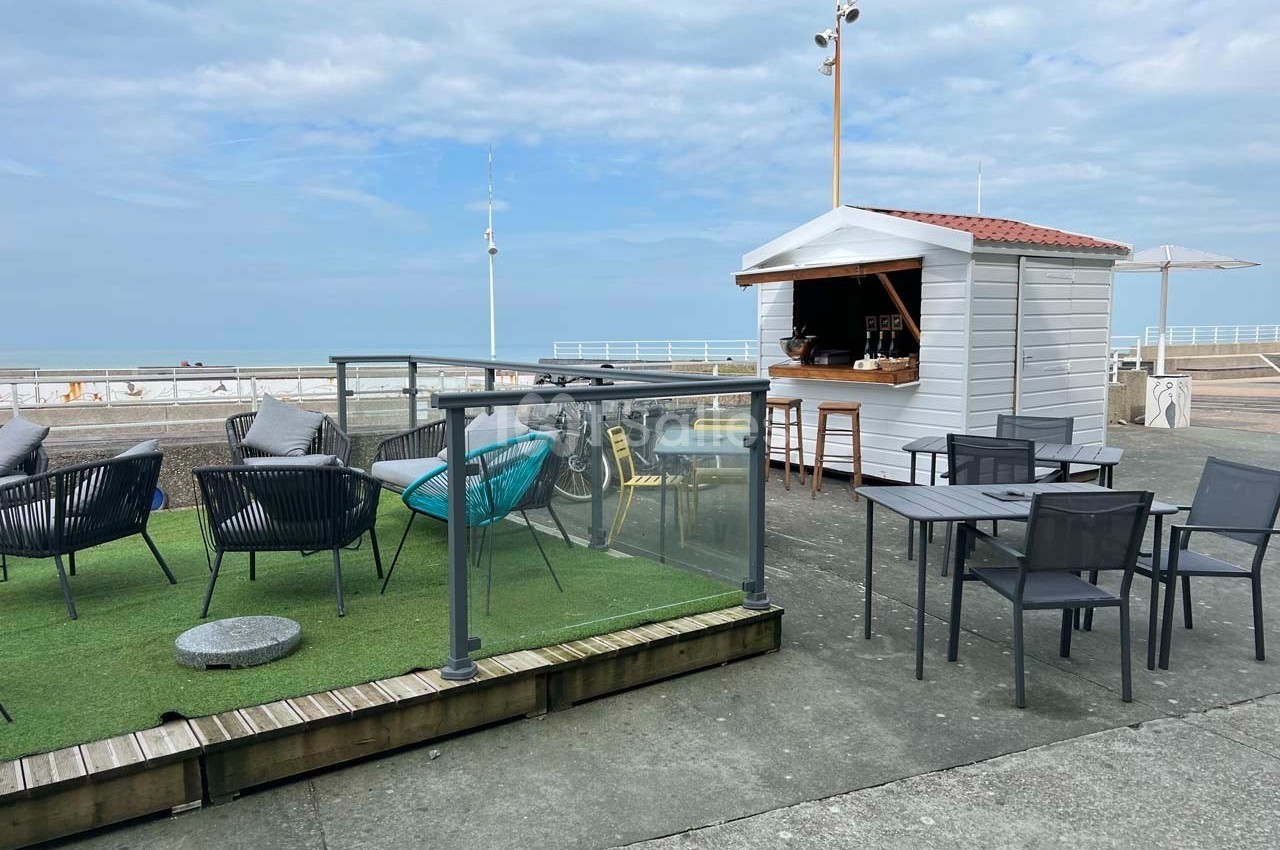 Terrasse en bord de mer avec chaises, tables, espace gazonné et un kiosque blanc sous un ciel partiellement nuageux.