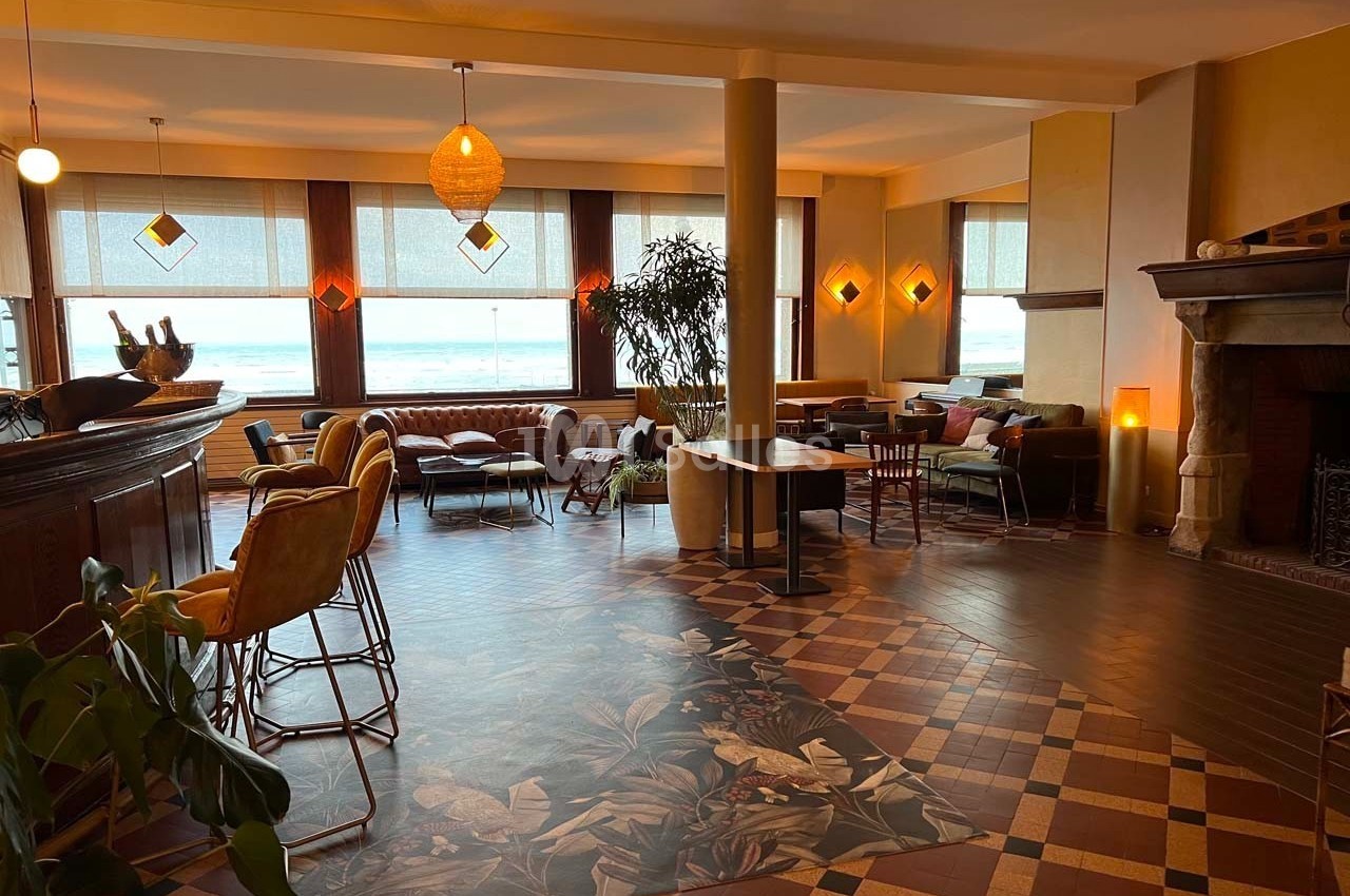 Salle lumineuse avec grandes fenêtres, mobilier varié, sol en carreaux décoratifs et cheminée en pierre.