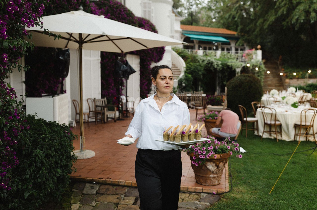 Une serveuse portant des verrines sur un plateau dans un jardin aménagé pour un événement en plein air.