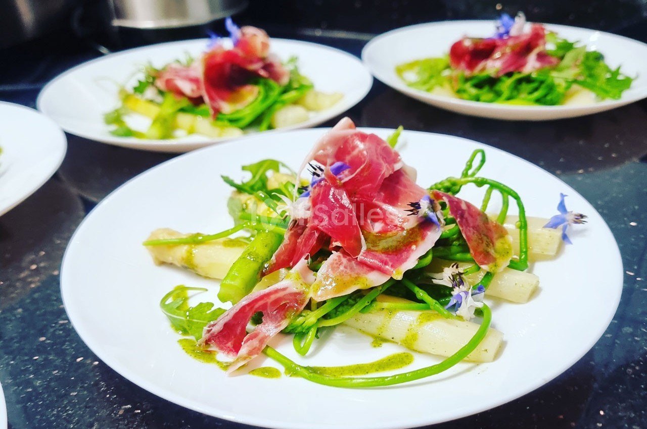 Assiette garnie d'asperges, roquette, jambon cru et fleurs comestibles, dressée sur une table sombre.