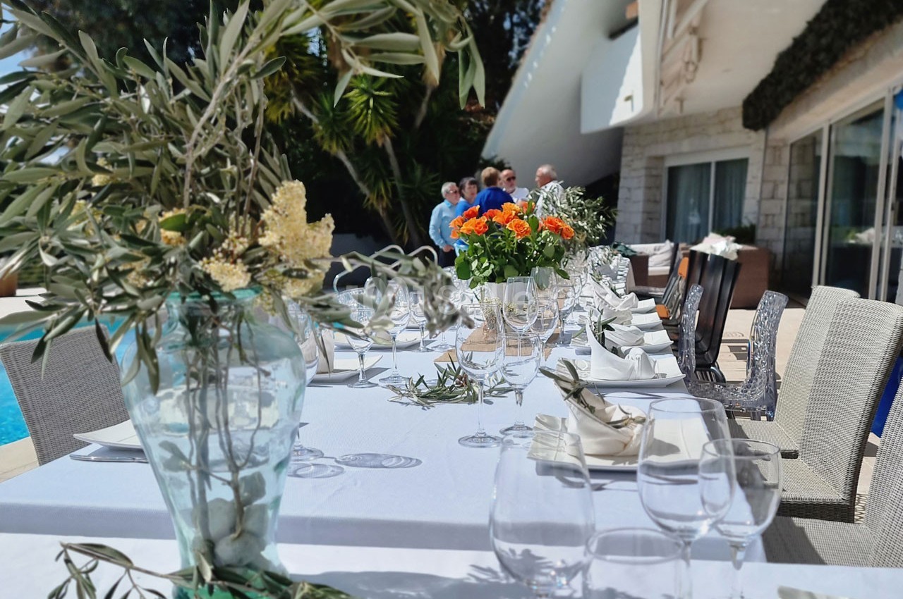 Table élégamment dressée en extérieur avec des fleurs et des verres, des convives discutant en arrière-plan.