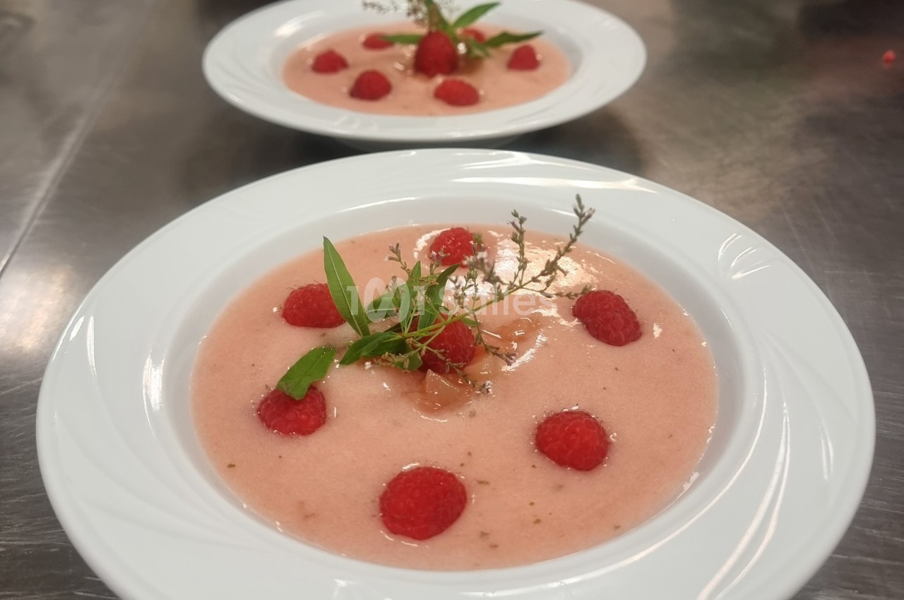 Deux assiettes de soupe froide rose garnies de framboises fraîches et de brins d'herbes aromatiques.